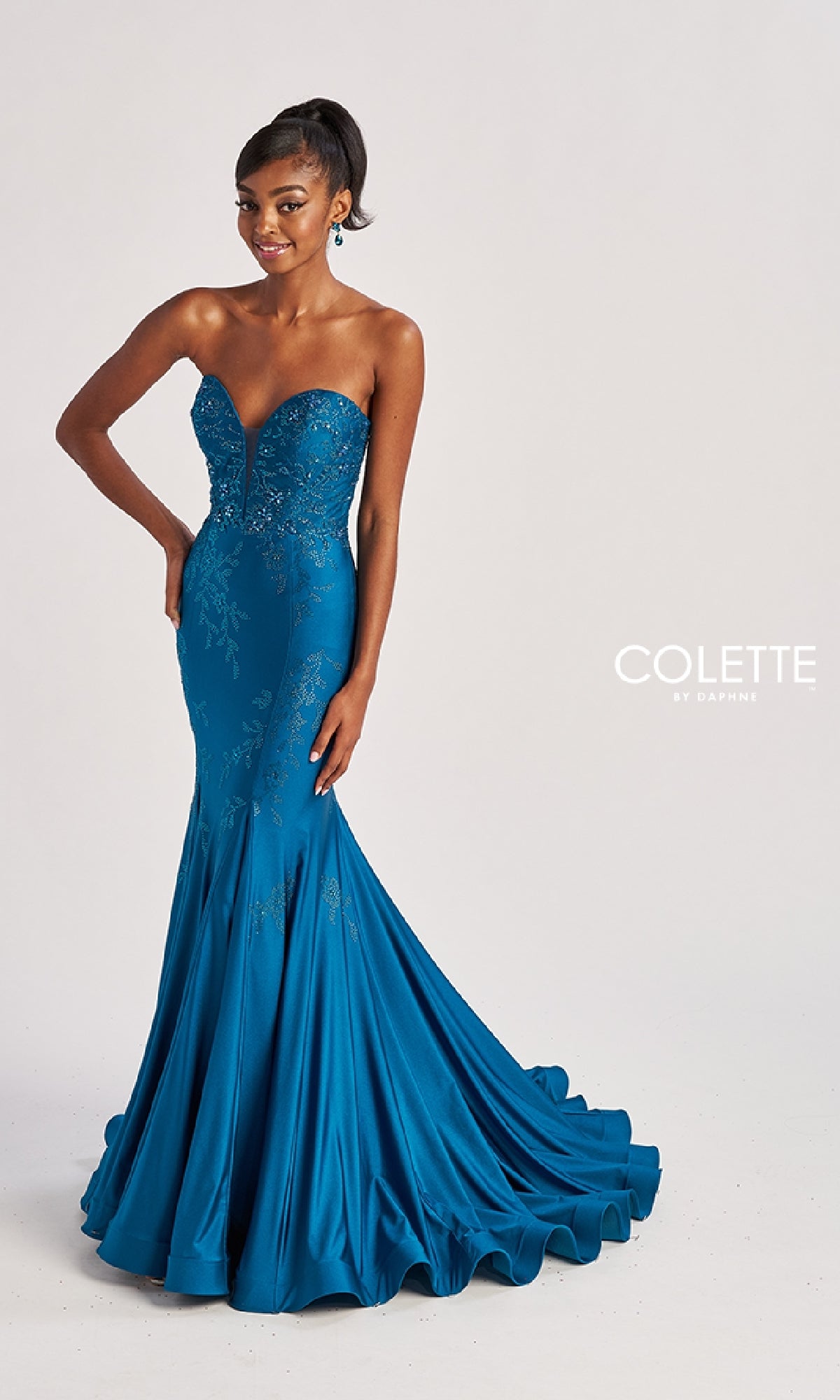 A woman stands against a plain background wearing the Colette CL8445, a floor-length teal strapless mermaid prom dress with a fitted silhouette, flared hem, subtle floral embellishments, and sweetheart neckline.